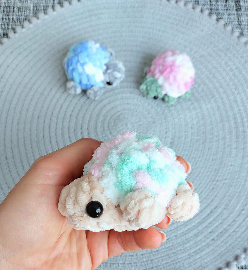 May include: Three small, plush, crocheted turtle figures. One is held in the foreground, featuring a beige head and limbs with a pastel pink, white, and mint green shell. Two smaller turtles in blue and pink sit in the background.
