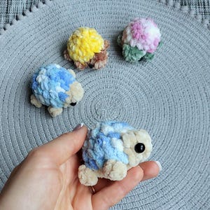 May include: Four small, handmade crochet plush turtles made from soft, textured yarn. One turtle is held in the foreground, while three others are arranged behind it. The turtles feature shells in yellow, pink and white, and blue and white, with beige heads and limbs.