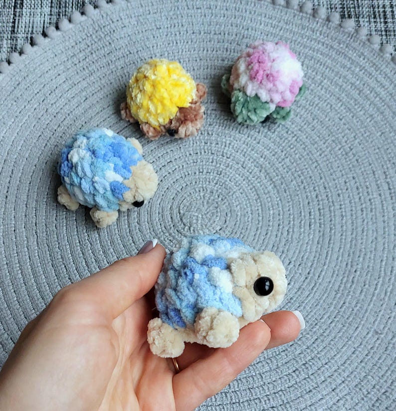 May include: Four small, handmade crochet plush turtles made from soft, textured yarn. One turtle is held in the foreground, while three others are arranged behind it. The turtles feature shells in yellow, pink and white, and blue and white, with beige heads and limbs.