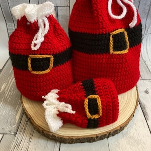 May include: Three crocheted Santa Claus gift bags in red, white, and black with a gold buckle. The bags are of different sizes and are sitting on a wooden surface.
