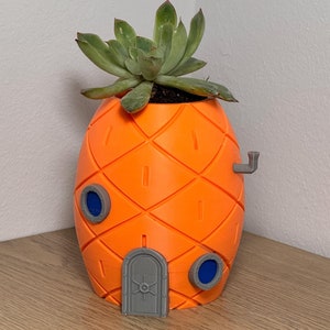 May include: Orange pineapple-shaped plant pot with a green succulent. The pot has a grey door, two blue porthole windows, and a grey chimney. The pineapple design features a diamond pattern with vertical rectangular cutouts.