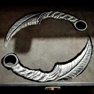 Puede incluir: Un par de cuchillos de lanzamiento decorativos en forma de pluma de color plateado con un anillo en el mango. Los cuchillos están dispuestos en círculo sobre una superficie gris oscuro.