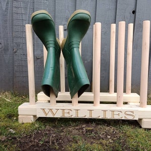 May include: Wooden boot rack with six slots for storing wellies. The rack is made of natural wood and has the word "WELLIES" engraved on the front.