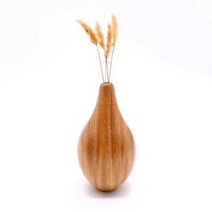 May include: A wooden vase with a teardrop shape, holding several dried, tan-coloured stems. The vase has a smooth, polished finish and a warm, brown colour. The vase is approximately 20 cm tall.