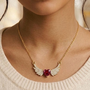 May include: A gold-toned necklace featuring a central heart-shaped red gemstone flanked by two outstretched wings encrusted with small clear crystals. The pendant is suspended from a delicate gold-toned chain that rests against the collarbone.