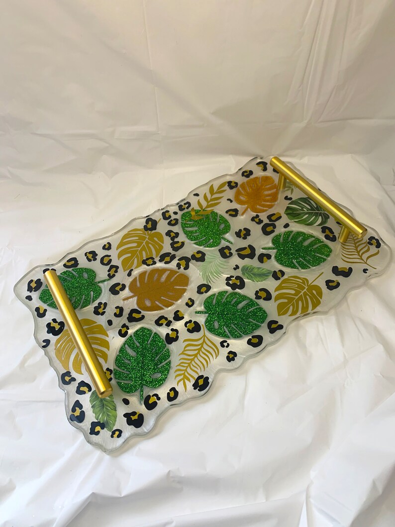 Puede incluir: Una bandeja de servir de resina transparente con asas doradas. La bandeja est&aacute; decorada con un estampado de leopardo y hojas de purpurina verde y dorada.