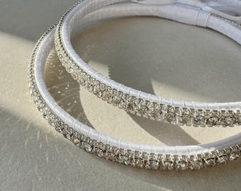 May include: Two white wedding crowns with a ribbon bow. Each crown is adorned with sparkling rhinestones and a satin ribbon. The crowns are circular and are designed for a wedding ceremony.