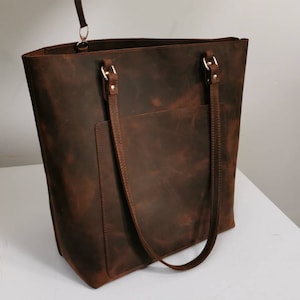 Könnte beinhalten: Eine braune Ledertasche mit einer Reißverschlusstasche. Die Tasche hat zwei Henkel und eine Fronttasche. Die Tasche hat einen goldenen Reißverschluss und einen Riemen.