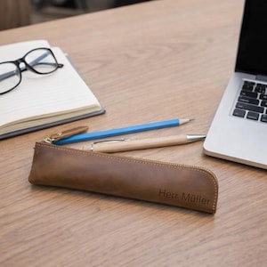 Op de afbeelding: Een bruine leren etui met de naam "Herr Müller" in reliëf, een blauw potlood en een houten pen liggen op een houten bureau. Een notitieboek met een bril en een laptop staan ook op het bureau.