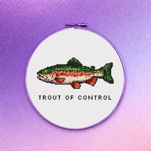 May include: A cross-stitch embroidery hoop featuring a pixelated rainbow trout design. The fish has green, red, orange, and white details. The text "TROUT OF CONTROL" is below the fish in black letters. The hoop is white with a purple border.