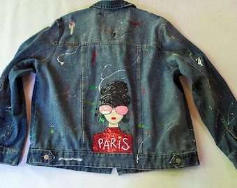 May include: A blue denim jacket with paint splatters and a hand-painted design on the back. The design features a woman with black hair, pink sunglasses, and a red shirt with the word "PARIS" written on it.