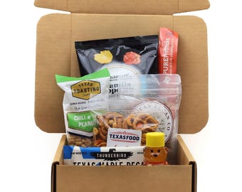 May include: A brown cardboard shipping box filled with various food items including a bag of Texas Roasting Company chili peanuts, a bag of pretzels, a Thunderbird Texas Maple Pecan bar, a small bear-shaped honey bottle, and other snack packages.