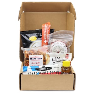 May include: A cardboard box filled with various snacks. The box contains bags of pretzels, nuts, and a bar. There is also a small bear-shaped bottle of honey and a packet of PureProSticks.