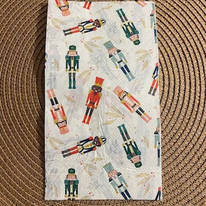 Op de afbeelding: Een witte papieren servet met een herhaald patroon van kleurrijke notenkrakerfiguren en gouden bladslingers. De notenkrakers zijn in verschillende kleuren, waaronder rood, groen, blauw en zwart, en zijn in verschillende richtingen gerangschikt.