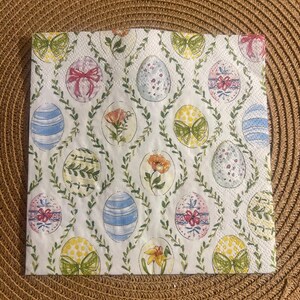 Pode incluir: Um guardanapo branco com tema de Páscoa com um padrão repetido de ovos decorados, flores e laços. Os ovos são adornados com listras, bolinhas e desenhos florais, rodeados por vinhas verdes. O guardanapo está sobre um sousplat marrom tecido.
