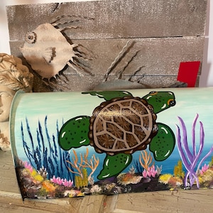 May include: A hand-painted mailbox featuring a sea turtle design. The turtle has a brown shell and green flippers. The background depicts an underwater scene with coral, seaweed, and a light blue sky. A metal fish sculpture and a bust are in the background.