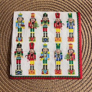 Op de afbeelding: Witte papieren servet met kleurrijke illustraties van speelgoednotenkrakers in verschillende uniformen. De notenkrakers zijn afgebeeld met gedetailleerde gezichtskenmerken, hoeden en accessoires. Het servet heeft een rode en groene rand, ideaal voor feestjes.