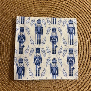 Peut inclure: Une serviette en papier blanche pliée avec un motif répété de soldats casse-noisettes bleus et de brins de feuilles bleues. Les casse-noisettes sont dans diverses poses, avec des uniformes détaillés.