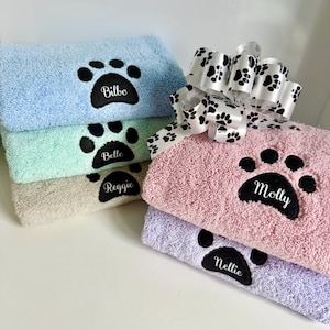 May include: A collection of pastel-coloured pet towels, including light blue, mint green, beige, pink, and lavender. Each towel has a black paw print with a pet's name embroidered in white. A decorative ribbon with paw prints is on top.