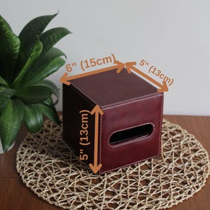 Puede incluir: Un cubre caja de pañuelos de cuero burdeos con una abertura cuadrada de 15 cm por 13 cm. La tapa mide 15 cm por 13 cm por 13 cm.