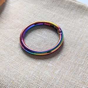 May include: A circular, iridescent metal clasp. The clasp is open, showing the hinge. The colours shift from red to blue and green. The clasp sits on a beige fabric surface.