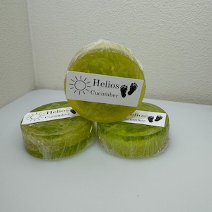 May include: Three round bars of green soap wrapped in plastic. Each bar has a white label with a sun and the text "Helios Cucumber".
