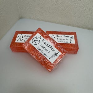 May include: Three orange soap bars with the text "Excalibur Leather & Labdanum" printed on them. The soap bars are wrapped in clear plastic.