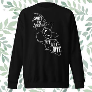 May include: Black sweatshirt featuring a white cartoon bat graphic. The bat has the text "SMALL AND SENSITIVE" above it and "BUT WILL BITE" below it. The sweatshirt is a long-sleeved crew neck.