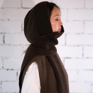 Könnte beinhalten: Dunkelbraunes Outfit, bestehend aus einem Kapuzenschal und einer Weste. Der Schal umschließt Hals und Kopf, mit einer Kapuze. Die Weste hat Taschen und wird über einem weißen Langarmhemd getragen.