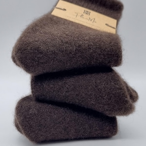 Può includere: Tre paia di calzini in alpaca marroni impilati uno sopra l'altro. La coppia in cima ha un piccolo cartellino marrone con il testo "Flan.Me" su di esso.