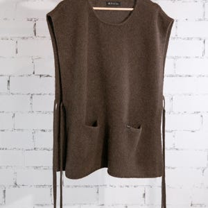 May include: A brown sleeveless sweater with two front pockets and side ties, hanging on a wooden hanger. The sweater has a round neckline and a textured knit. The background is a white brick wall.