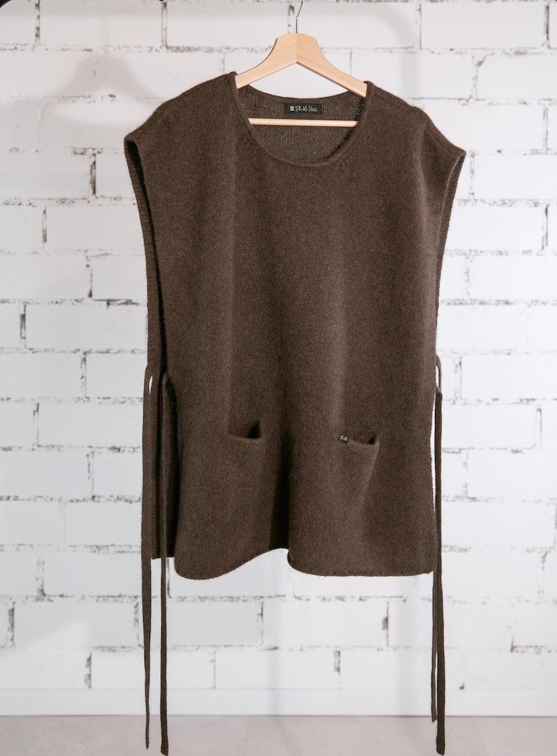 May include: A brown sleeveless sweater with two front pockets and side ties, hanging on a wooden hanger. The sweater has a round neckline and a textured knit. The background is a white brick wall.