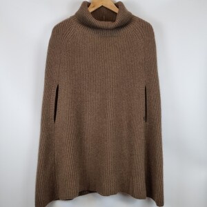 May include: A brown, ribbed knit poncho featuring a high turtleneck collar and vertical knit patterns. The garment includes two side arm openings and a flared silhouette, providing a structured yet relaxed fit for layering over various outfits.