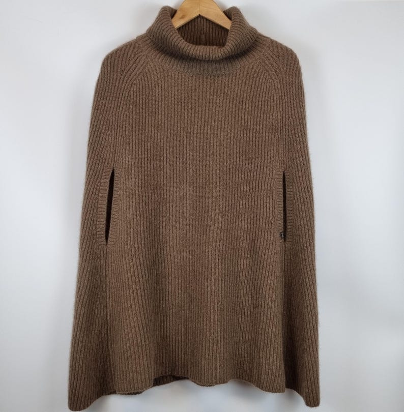 May include: A brown, ribbed knit poncho featuring a high turtleneck collar and vertical knit patterns. The garment includes two side arm openings and a flared silhouette, providing a structured yet relaxed fit for layering over various outfits.