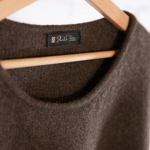 May include: A close-up of a brown sweater with a crew neck, hanging on a wooden hanger. The sweater has a tag that reads "Plan Me" in white text on a black background. The sweater's texture is visible.