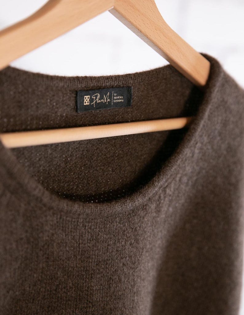 May include: A close-up of a brown sweater with a crew neck, hanging on a wooden hanger. The sweater has a tag that reads "Plan Me" in white text on a black background. The sweater's texture is visible.