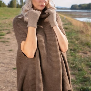 May include: A brown ribbed knit poncho with a high collar, paired with matching light brown knit gloves and a dark brown felt cap. The poncho features a sleeveless design with wide arm openings and a vertical ribbed texture throughout the fabric.