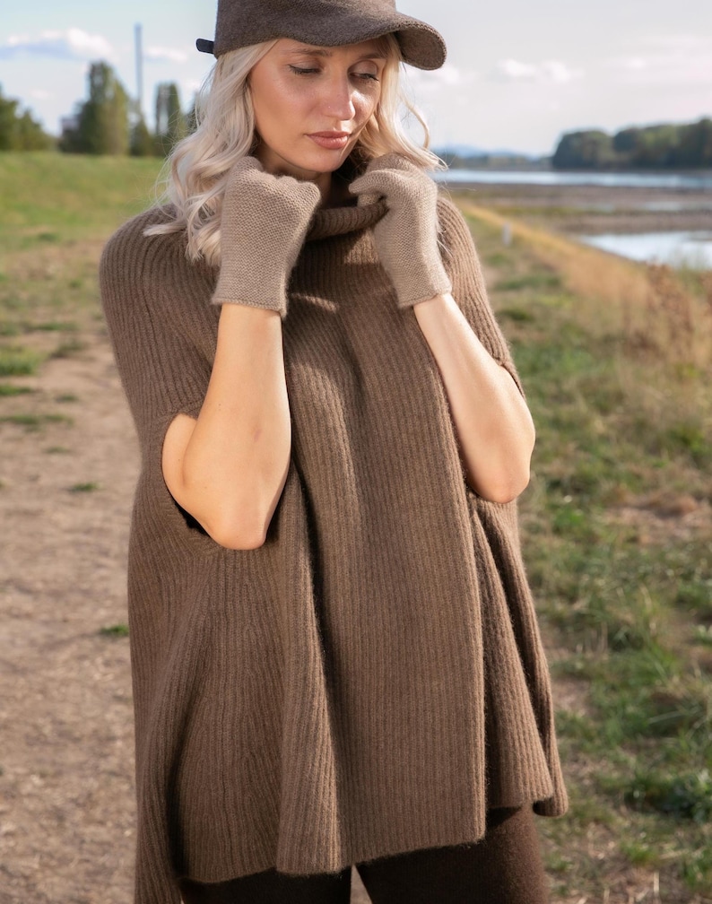 May include: A brown ribbed knit poncho with a high collar, paired with matching light brown knit gloves and a dark brown felt cap. The poncho features a sleeveless design with wide arm openings and a vertical ribbed texture throughout the fabric.