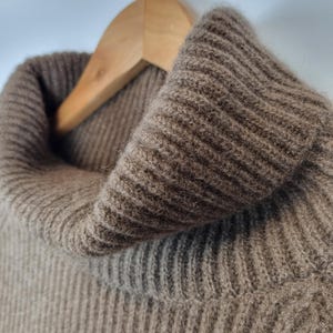 May include: A brown turtleneck sweater featuring a thick, ribbed knit texture. The garment is constructed with a prominent, folded collar that extends upward, showcasing the detailed vertical ribbing pattern throughout the fabric surface.
