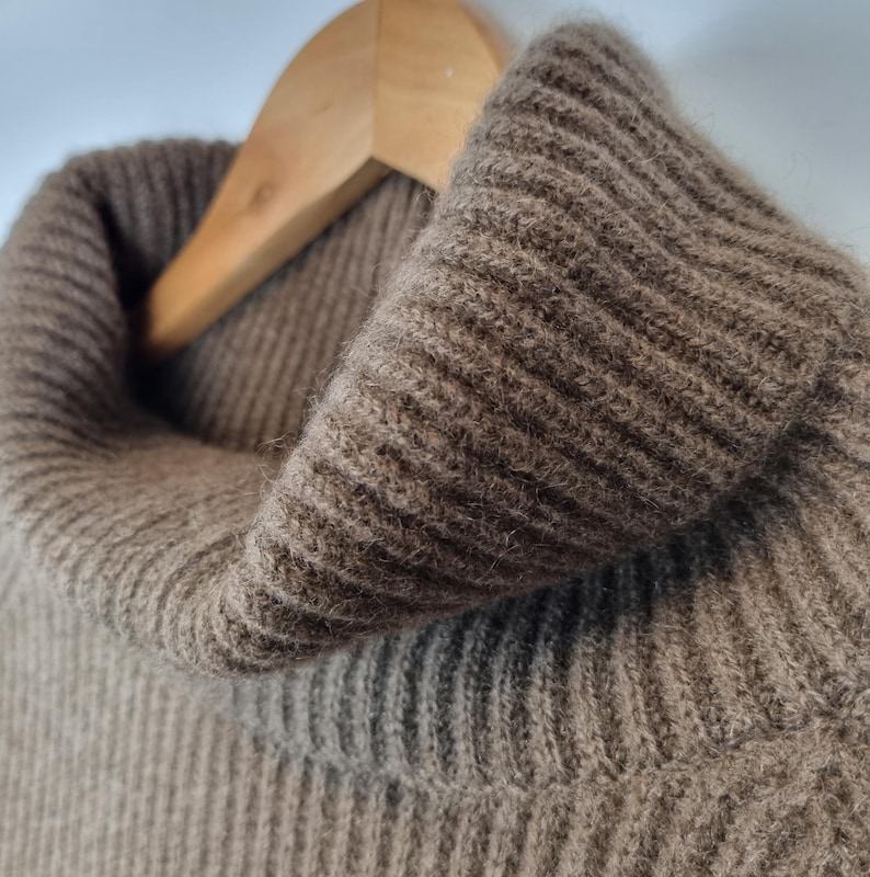 May include: A brown turtleneck sweater featuring a thick, ribbed knit texture. The garment is constructed with a prominent, folded collar that extends upward, showcasing the detailed vertical ribbing pattern throughout the fabric surface.