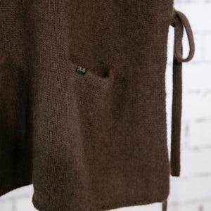 May include: Close-up of a dark brown knitted garment with a small pocket and a tie closure. The fabric has a textured appearance. A small label is sewn on the pocket. The garment appears to be a vest or wrap.