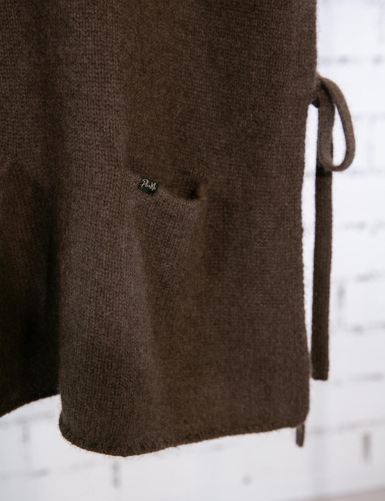 May include: Close-up of a dark brown knitted garment with a small pocket and a tie closure. The fabric has a textured appearance. A small label is sewn on the pocket. The garment appears to be a vest or wrap.