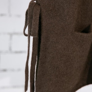 May include: Close-up of a brown knit vest with a side tie and a pocket. The vest is made of a soft, textured material. The tie is made of the same material as the vest. The background is a white brick wall.