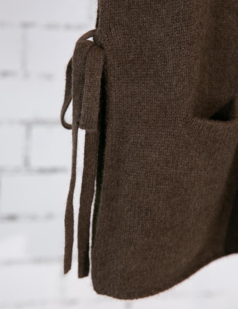May include: Close-up of a brown knit vest with a side tie and a pocket. The vest is made of a soft, textured material. The tie is made of the same material as the vest. The background is a white brick wall.
