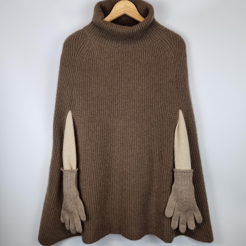 May include: A brown ribbed knit poncho featuring a high turtleneck collar. The garment includes two beige fabric sleeves extending from the side openings, each finished with a pair of matching brown knit gloves attached at the wrist area.