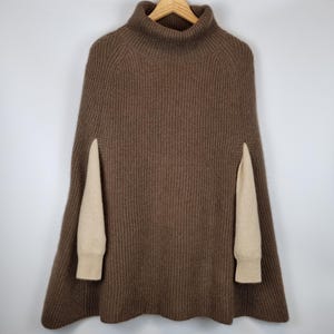 May include: A brown ribbed knit poncho featuring a high turtleneck collar and wide arm openings. The garment displays a textured vertical knit pattern throughout the body, with contrasting beige sleeves visible through the side slits of the poncho.