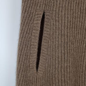 May include: A brown knit garment featuring a vertical ribbed texture and a single integrated slit pocket. The fabric has a soft, fuzzy appearance with a consistent weave pattern throughout the piece, highlighting the detailed construction of the pocket opening.