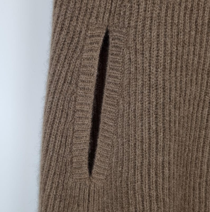 May include: A brown knit garment featuring a vertical ribbed texture and a single integrated slit pocket. The fabric has a soft, fuzzy appearance with a consistent weave pattern throughout the piece, highlighting the detailed construction of the pocket opening.