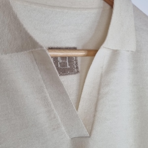 May include: An off-white knit polo shirt featuring a V-neckline with a collar and a small, square, grey-toned fabric label stitched inside the neck area. The garment is displayed on a wooden hanger, showcasing the fine texture of the knit fabric.