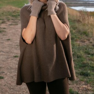 May include: A brown ribbed knit poncho with a high turtleneck collar and matching brown knit gloves. The garment features an open-side design with arm openings, providing a loose, flowing fit that extends to the upper thighs in a solid earth tone.